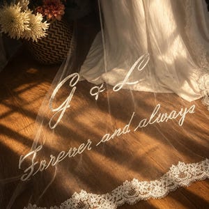 Könnte beinhalten: Ein weißer Brautschleier aus Tüll mit einer aufwendigen floralen Spitzenborte am Rand. Der Schleier ist mit dem weißen Schriftzug G & L und dem Satz Forever and always bestickt. Der Stoff ist transparent und liegt auf einem Holzboden.