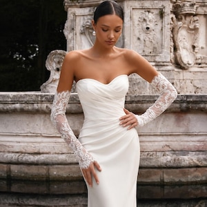 May include: A strapless white floor-length gown featuring a sweetheart neckline with diagonal ruched fabric across the bodice and fitted skirt. The garment is paired with long, sheer white lace gloves that extend from the wrists to the upper arms.
