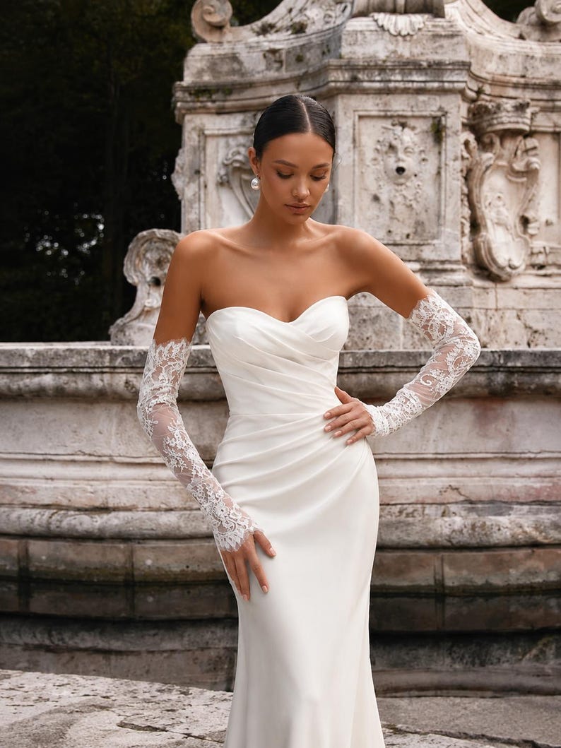 May include: A strapless white floor-length gown featuring a sweetheart neckline with diagonal ruched fabric across the bodice and fitted skirt. The garment is paired with long, sheer white lace gloves that extend from the wrists to the upper arms.
