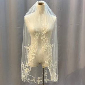 May include: A white bridal veil with delicate floral embroidery and pearl accents. The veil is displayed on a mannequin.