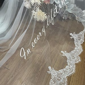 May include: A white bridal veil with delicate lace trim and the phrase "In every lifetime" in elegant script. The veil is draped over a wooden surface, with a basket of flowers in the background.