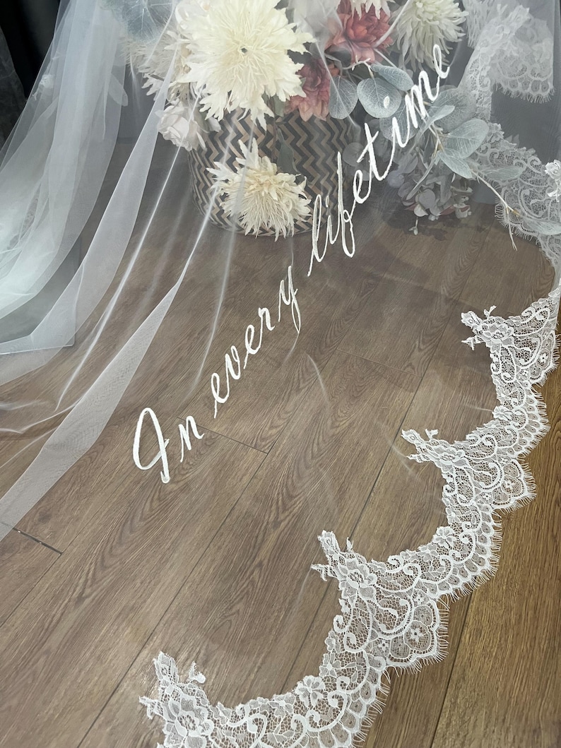 May include: A white bridal veil with delicate lace trim and the phrase "In every lifetime" in elegant script. The veil is draped over a wooden surface, with a basket of flowers in the background.