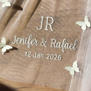 May include: A sheer, beige fabric with white embroidered text reading "JR, Jenifer & Rafael, 12 Jan 2026." Several white butterfly appliqu&eacute;s are scattered across the fabric, creating a delicate, romantic design.