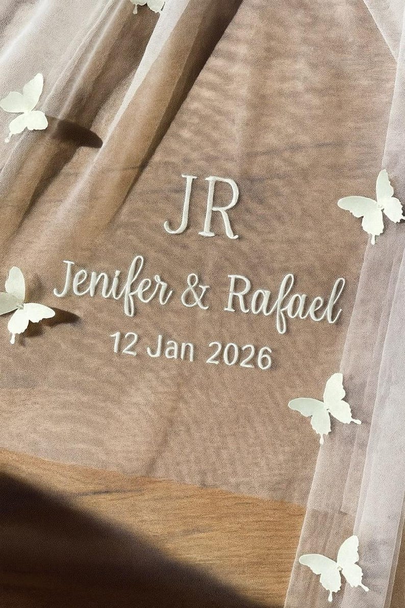 May include: A sheer, beige fabric with white embroidered text reading "JR, Jenifer & Rafael, 12 Jan 2026." Several white butterfly appliqu&eacute;s are scattered across the fabric, creating a delicate, romantic design.