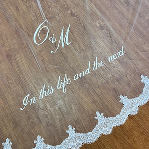 以下が含まれることがあります： 繊細なレーストリムが付いた、透明感のある白いウェディングベール。「O&M」のイニシャルと「In this life and the next」というフレーズがエレガントな筆記体で書かれており、花嫁に最適です。