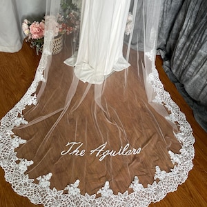 May include: A white wedding veil with a delicate lace trim and the embroidered text "The Aguilars". The veil is draped over a white dress, with a wooden floor in the background. The lace trim is detailed and intricate.