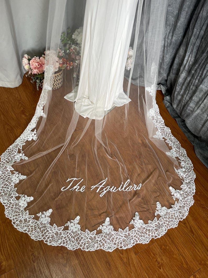 May include: A white wedding veil with a delicate lace trim and the embroidered text "The Aguilars". The veil is draped over a white dress, with a wooden floor in the background. The lace trim is detailed and intricate.