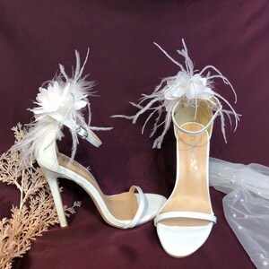 May include: White bridal heels with a feather and flower embellishment. The heels are made of satin and have a delicate strap around the ankle.