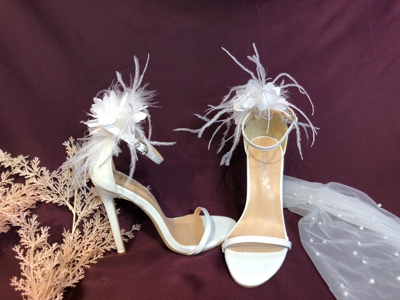 May include: White bridal heels with a feather and flower embellishment. The heels are made of satin and have a delicate strap around the ankle.