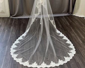 May include: A long, sheer white bridal veil with a delicate, scalloped lace trim. The lace trim is adorned with small, scattered pearls. The veil is displayed against a neutral background, showcasing its elegant design and texture.