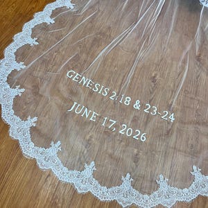 May include: A sheer white wedding veil with a delicate lace trim along the edge. The text "GENESIS 2:18 & 23-24" and "JUNE 17, 2026" are printed in white on the veil. The veil is draped over a wooden surface.
