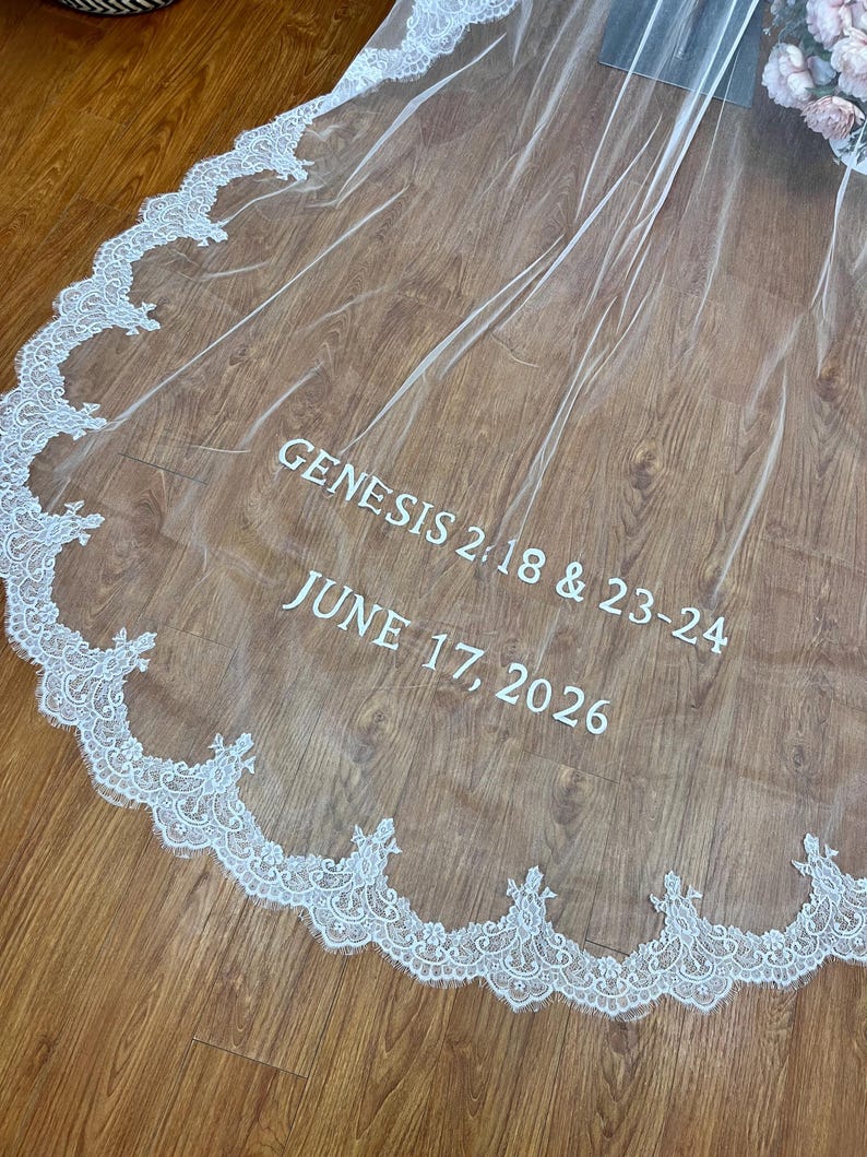 May include: A sheer white wedding veil with a delicate lace trim along the edge. The text "GENESIS 2:18 & 23-24" and "JUNE 17, 2026" are printed in white on the veil. The veil is draped over a wooden surface.