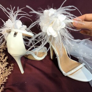 May include: White bridal heels with a large white flower and feather embellishment. The heels have a delicate ankle strap and a high stiletto heel.
