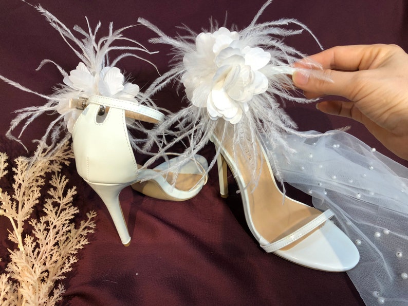 May include: White bridal heels with a large white flower and feather embellishment. The heels have a delicate ankle strap and a high stiletto heel.
