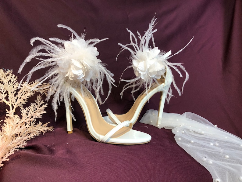 May include: White high-heeled sandals with a large white flower and feather embellishment on each strap.