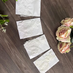 May include: Four white lace fabric samples on white cards, each with a different intricate floral design. The samples are labeled "White", "Off White", and "Light Ivory". The samples are arranged on a dark wood surface, with pink roses and greenery.