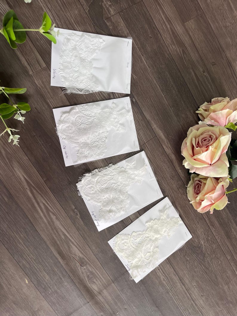 May include: Four white lace fabric samples on white cards, each with a different intricate floral design. The samples are labeled "White", "Off White", and "Light Ivory". The samples are arranged on a dark wood surface, with pink roses and greenery.