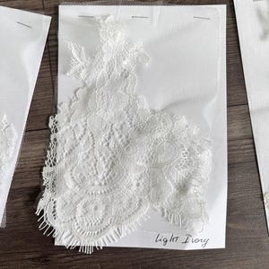 May include: Close-up of light ivory lace fabric samples. The lace features intricate floral and geometric patterns. The samples are attached to white backing material and are labeled "Light Ivory".