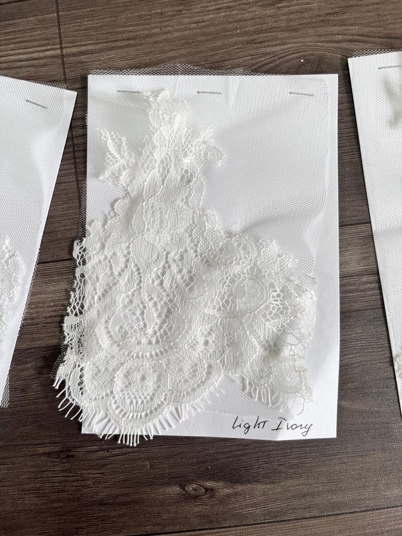 May include: Close-up of light ivory lace fabric samples. The lace features intricate floral and geometric patterns. The samples are attached to white backing material and are labeled "Light Ivory".