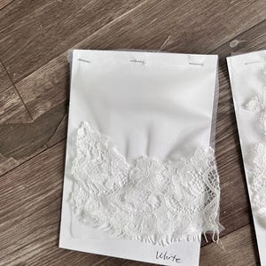 May include: White lace fabric samples attached to white cards with the word "White" written on them. The lace has a delicate floral pattern and is stapled to a sheer backing. The samples are displayed on a wooden surface.