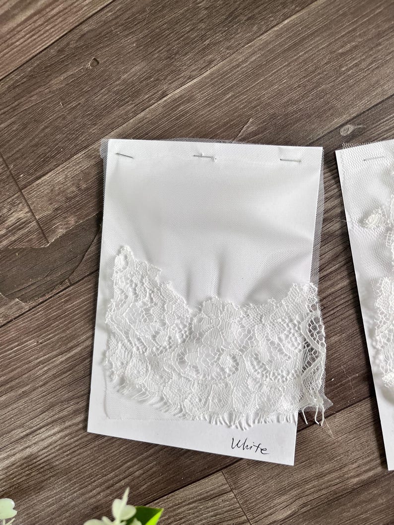 May include: White lace fabric samples attached to white cards with the word "White" written on them. The lace has a delicate floral pattern and is stapled to a sheer backing. The samples are displayed on a wooden surface.