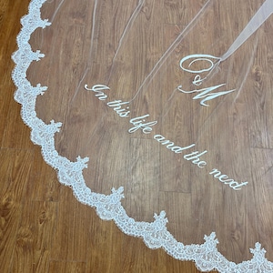 May include: A sheer white wedding veil with a delicate lace trim along the edge. The veil features embroidered text that reads "In this life and the next" and initials "C & M". The veil is displayed on a wooden surface.