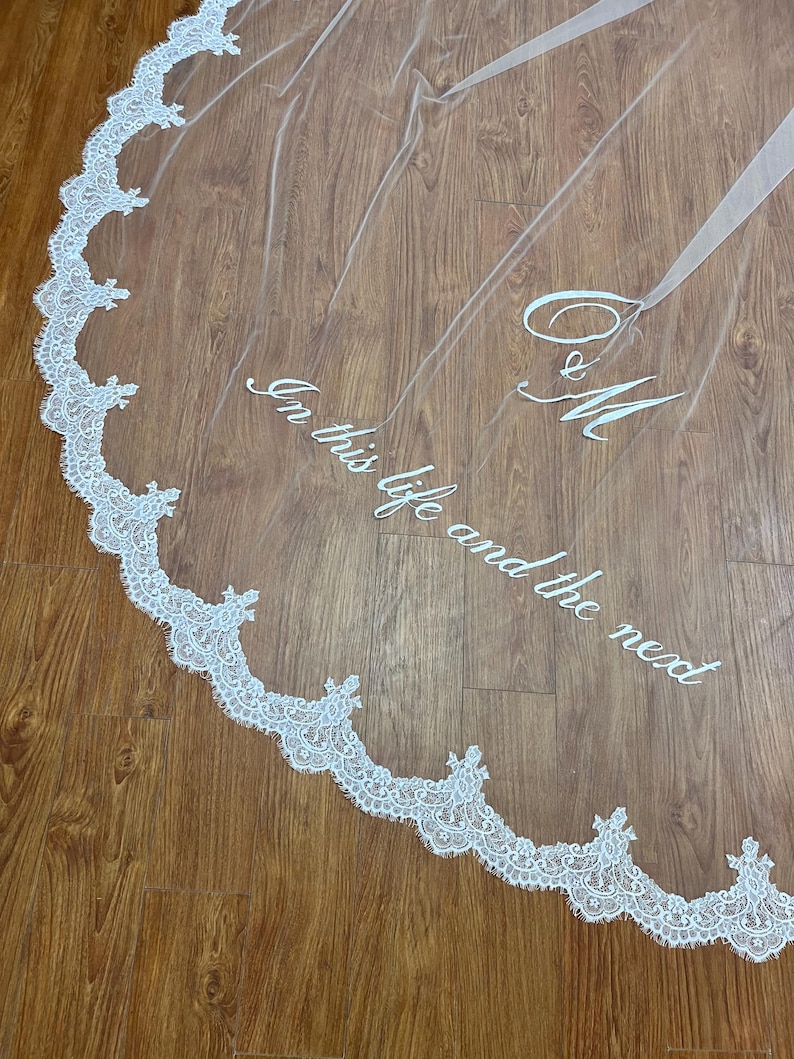 May include: A sheer white wedding veil with a delicate lace trim along the edge. The veil features embroidered text that reads "In this life and the next" and initials "C & M". The veil is displayed on a wooden surface.
