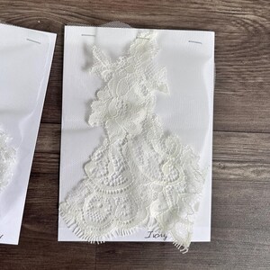 May include: Ivory lace applique on white fabric, secured with pins. The delicate floral lace design is displayed against a white background. The word "Ivory" is handwritten in cursive. Ideal for bridal or craft projects.