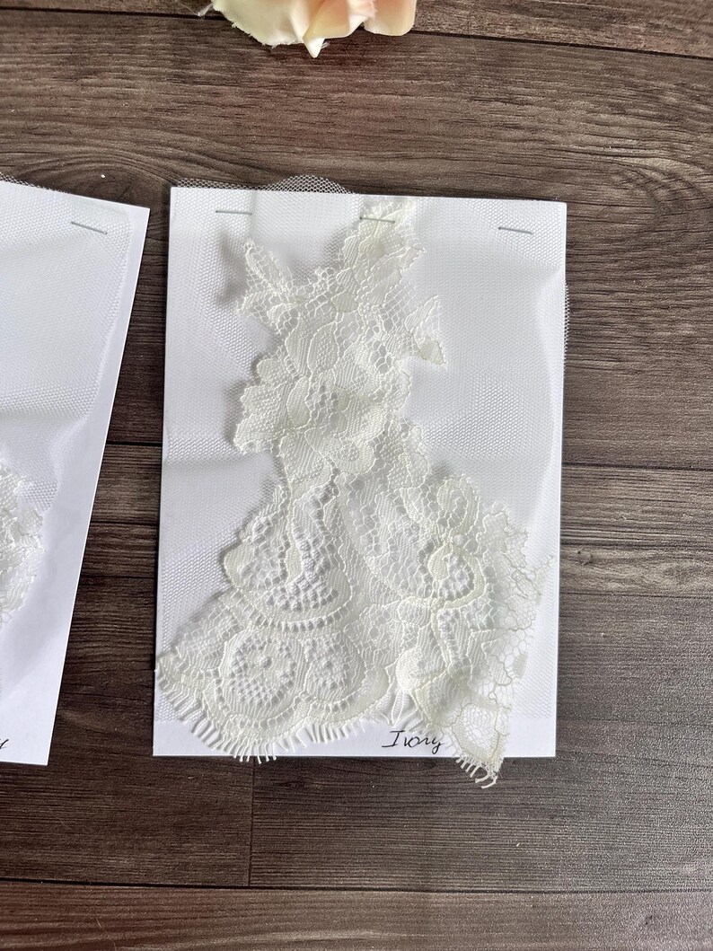 May include: Ivory lace applique on white fabric, secured with pins. The delicate floral lace design is displayed against a white background. The word "Ivory" is handwritten in cursive. Ideal for bridal or craft projects.