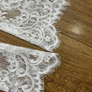 May include: A pair of white lace sleeves featuring an intricate floral pattern with scalloped edges and delicate fringe detailing. The fabric is sheer with a fine mesh background, showcasing a detailed botanical design throughout the material.