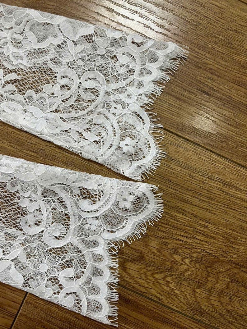 May include: A pair of white lace sleeves featuring an intricate floral pattern with scalloped edges and delicate fringe detailing. The fabric is sheer with a fine mesh background, showcasing a detailed botanical design throughout the material.
