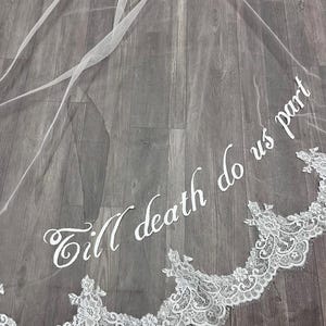 May include: A white bridal veil with delicate lace trim along the bottom edge. The veil features the embroidered phrase "Till death do us part" in elegant script, adding a romantic touch to the wedding accessory.
