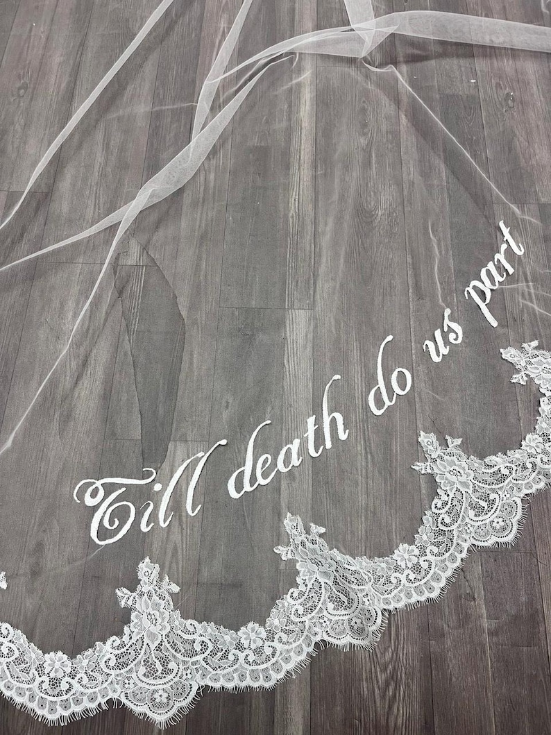 May include: A white bridal veil with delicate lace trim along the bottom edge. The veil features the embroidered phrase "Till death do us part" in elegant script, adding a romantic touch to the wedding accessory.