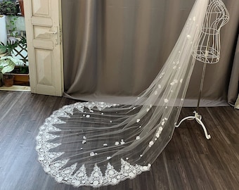 May include: A white bridal veil with a delicate floral lace trim and scattered 3D flowers. The veil is made of soft tulle and is designed to drape elegantly over the bride's shoulders.