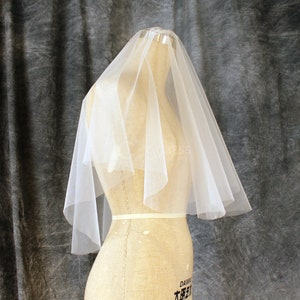 Puede incluir: Un velo de novia de tul blanco de una sola capa con un diseño sencillo. El velo se muestra en un maniquí.