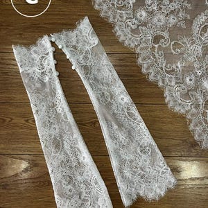 May include: A pair of white lace fingerless gloves featuring intricate floral patterns and scalloped edges, with a row of small round buttons along the side. A matching piece of white lace fabric with a scalloped border is positioned nearby.