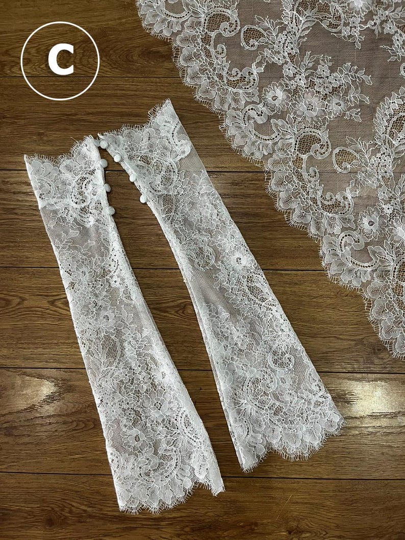 May include: A pair of white lace fingerless gloves featuring intricate floral patterns and scalloped edges, with a row of small round buttons along the side. A matching piece of white lace fabric with a scalloped border is positioned nearby.
