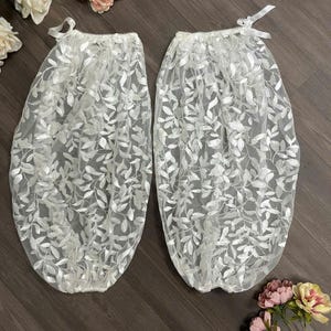 May include: Two white, leaf-patterned lace sleeves with a delicate, sheer appearance. Each sleeve is gathered at the top and tied with a small white ribbon. The sleeves are displayed on a dark wood surface, with floral accents.