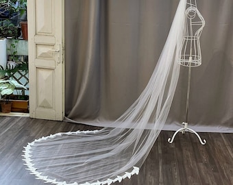 May include: A white tulle wedding veil with a delicate lace trim. The veil is draped over a white mannequin and is displayed on a wooden floor.
