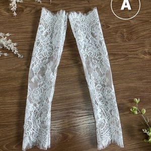 May include: A pair of white lace arm sleeves featuring a delicate floral pattern and scalloped edges at the top and bottom openings. The sheer fabric displays an intricate, detailed design throughout the length of each sleeve, providing a textured look.