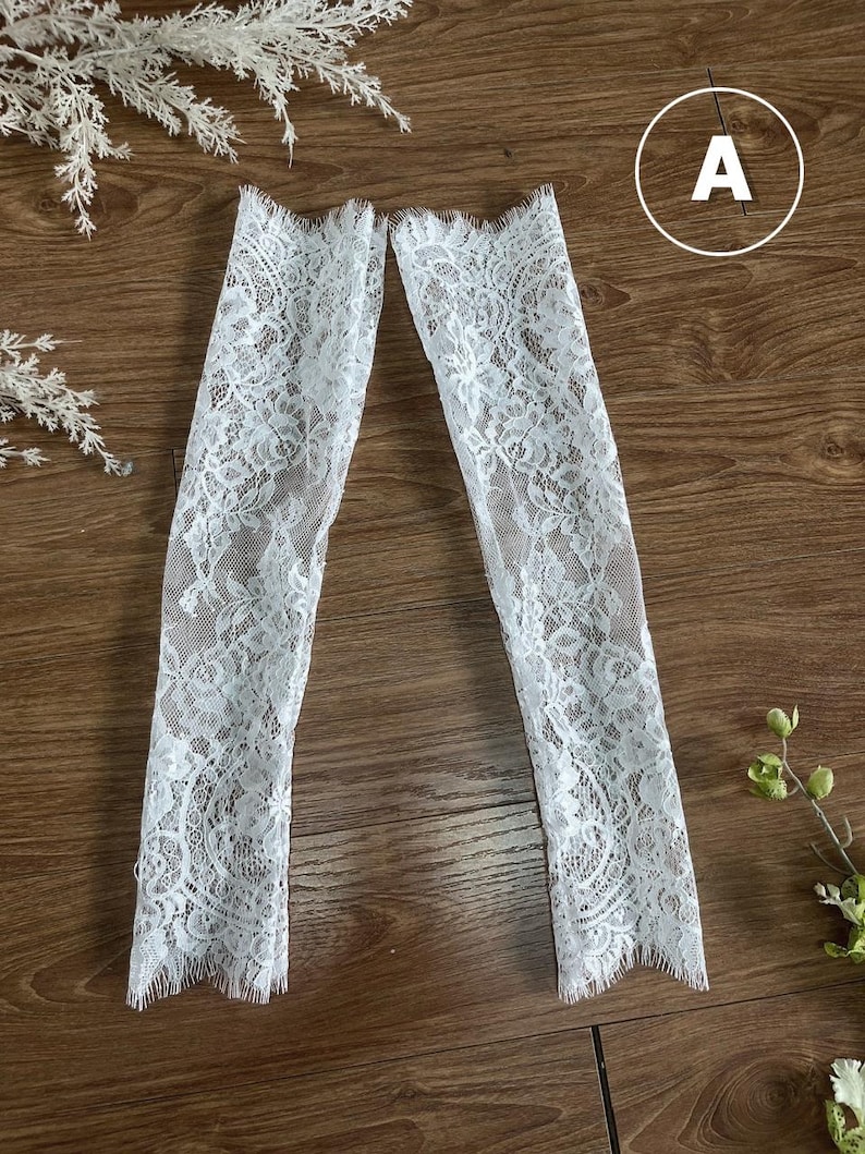 May include: A pair of white lace arm sleeves featuring a delicate floral pattern and scalloped edges at the top and bottom openings. The sheer fabric displays an intricate, detailed design throughout the length of each sleeve, providing a textured look.