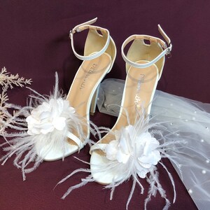 May include: White bridal heels with a feather and flower embellishment. The heels have a delicate ankle strap and a high stiletto heel.