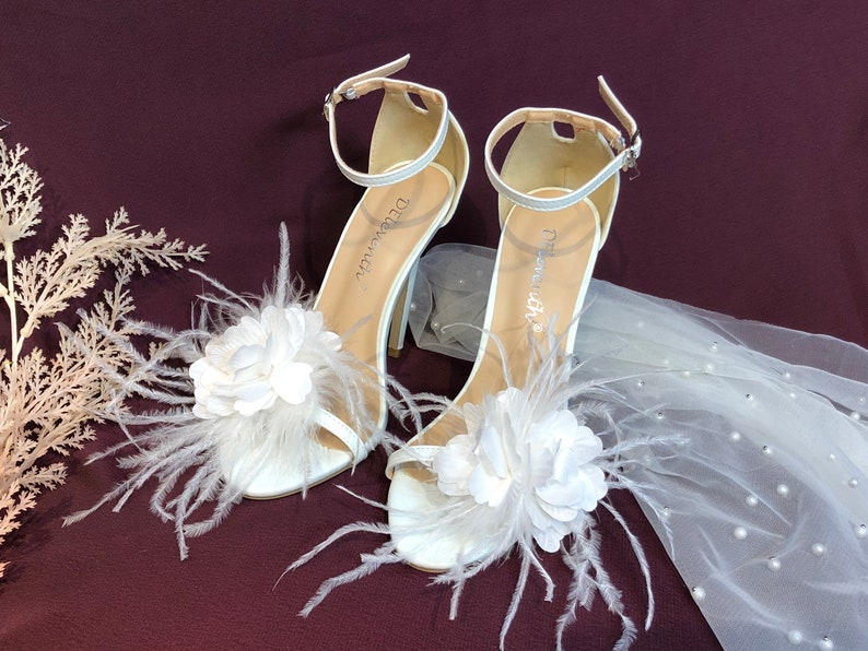 May include: White bridal heels with a feather and flower embellishment. The heels have a delicate ankle strap and a high stiletto heel.