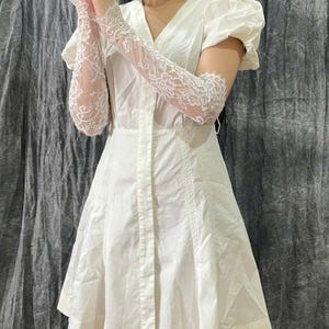 May include: A white short-sleeved dress featuring a V-neckline, a button-down front, and a flared skirt, paired with long, sheer white lace gloves that extend past the elbows, showcasing intricate floral patterns and scalloped edges.