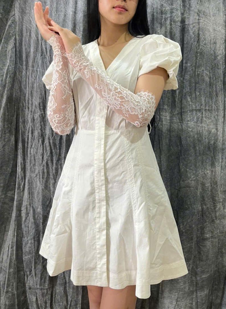 May include: A white short-sleeved dress featuring a V-neckline, a button-down front, and a flared skirt, paired with long, sheer white lace gloves that extend past the elbows, showcasing intricate floral patterns and scalloped edges.