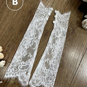 May include: A pair of white lace fingerless gloves featuring a floral pattern with scalloped edges and three small white buttons along the side of each cuff. The letter B is visible in a white circle in the upper left corner of the image.