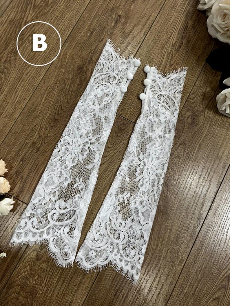 May include: A pair of white lace fingerless gloves featuring a floral pattern with scalloped edges and three small white buttons along the side of each cuff. The letter B is visible in a white circle in the upper left corner of the image.