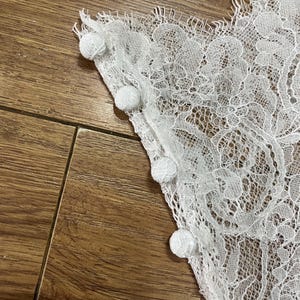 May include: A white lace fabric panel featuring a delicate floral pattern with scalloped edges and a row of four small, round, fabric-covered buttons attached along the straight vertical side seam of the garment piece.