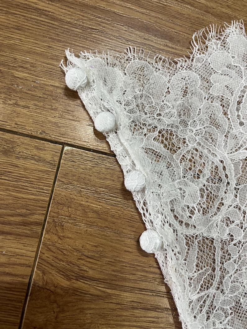 May include: A white lace fabric panel featuring a delicate floral pattern with scalloped edges and a row of four small, round, fabric-covered buttons attached along the straight vertical side seam of the garment piece.