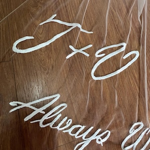 May include: A sheer white bridal veil with embroidered white text. The text includes the initials "J x C" and the words "Always & Forever". The veil is draped over a wooden surface.